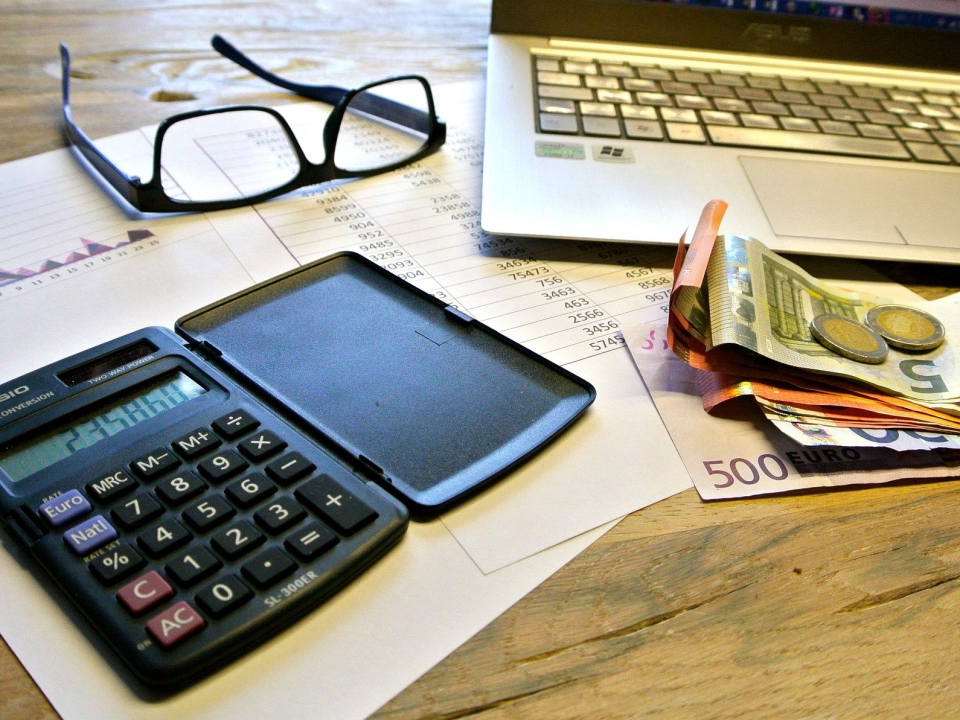 Brille, Taschenrechner, Laptop und Geld liegen gemeinsam auf einem Schreibtisch